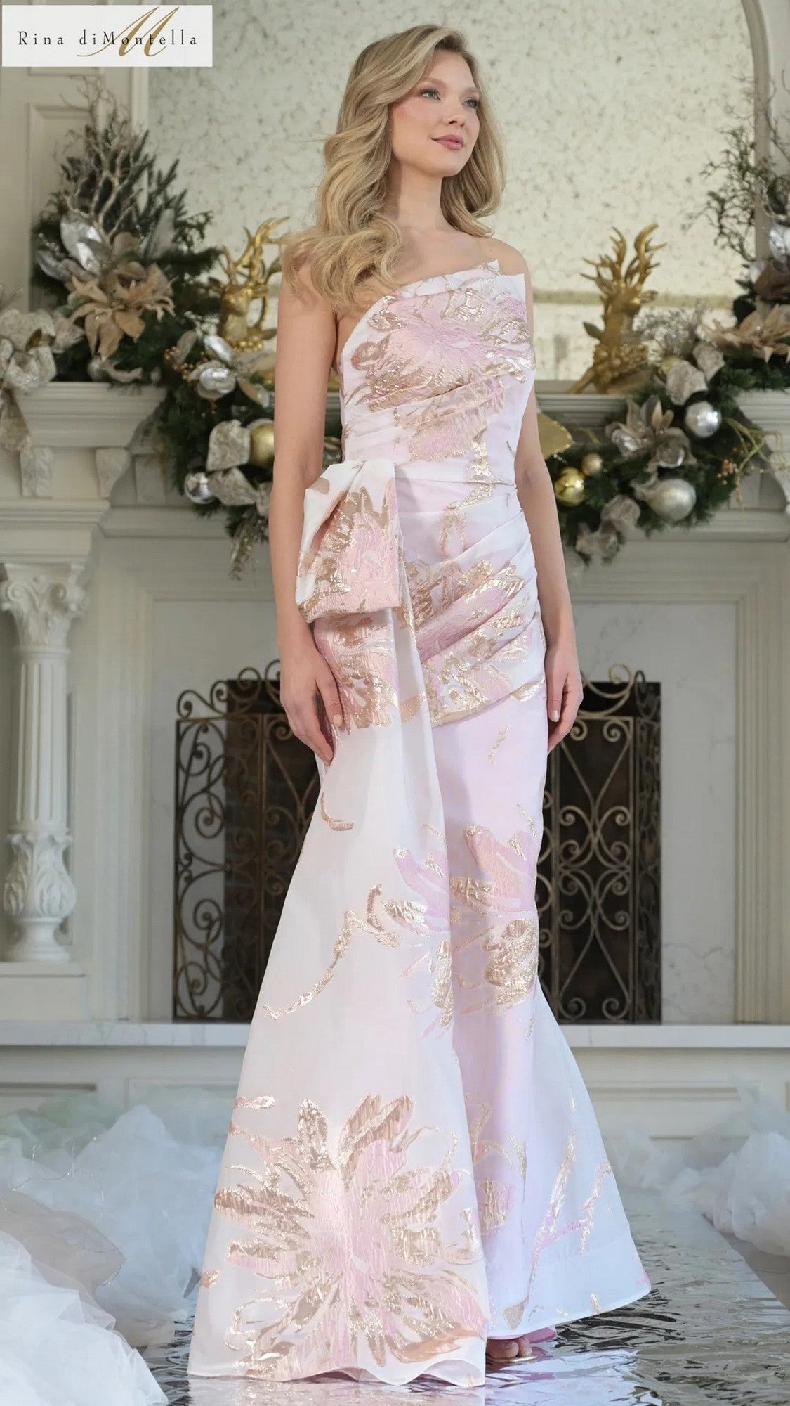 A woman indoors wears the Rina Di Montella RD3233 long formal dress in blush multi, featuring a floral organza jacquard with metallic patterns and a statement side bow, surrounded by holiday decorations.
